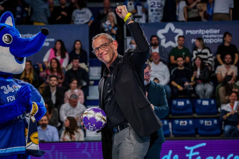 Homme souriant salue public avec mascotte sportive.